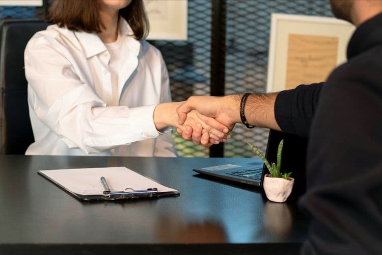 Candidate and interviewer shaking hands across a desk after a successful job interview, with CV clipboard and laptop visible