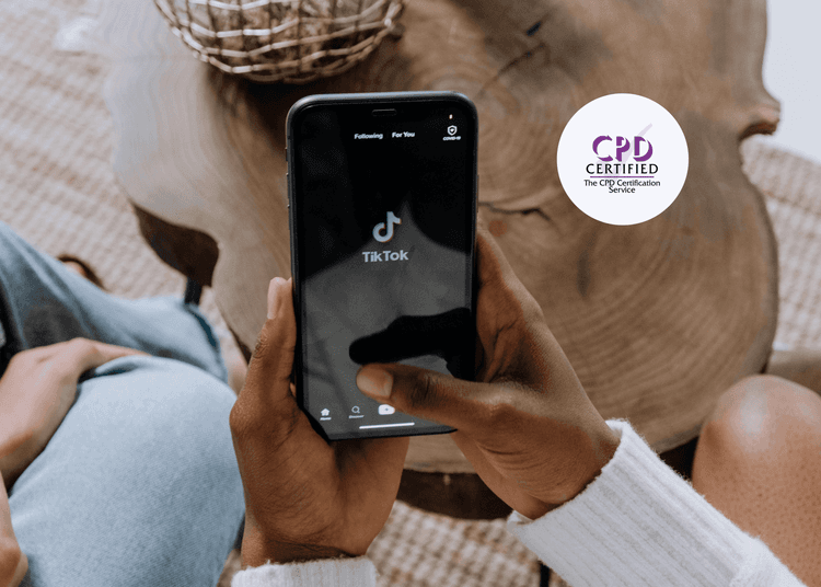 A TikTok launch screen shown on a mobile device, supporting CPD learning on youth engagement with social media platforms. Close-up of hands holding a smartphone displaying the TikTok app home screen, with two people seated on a wooden table.